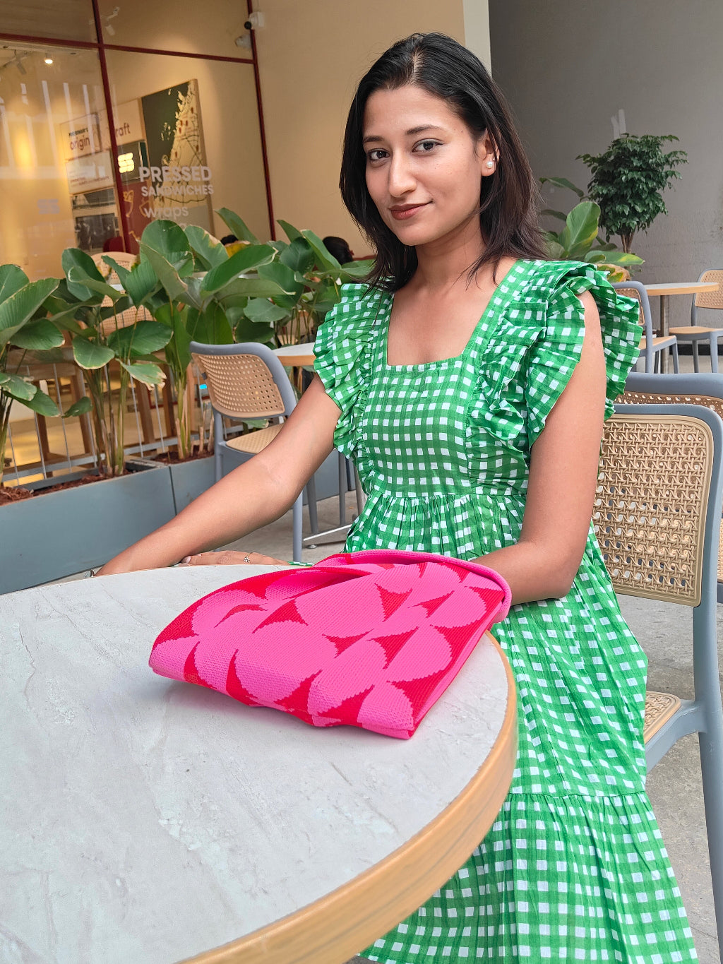 Woman in a green checkered dress holding a pink floral bag in an indoor setting.